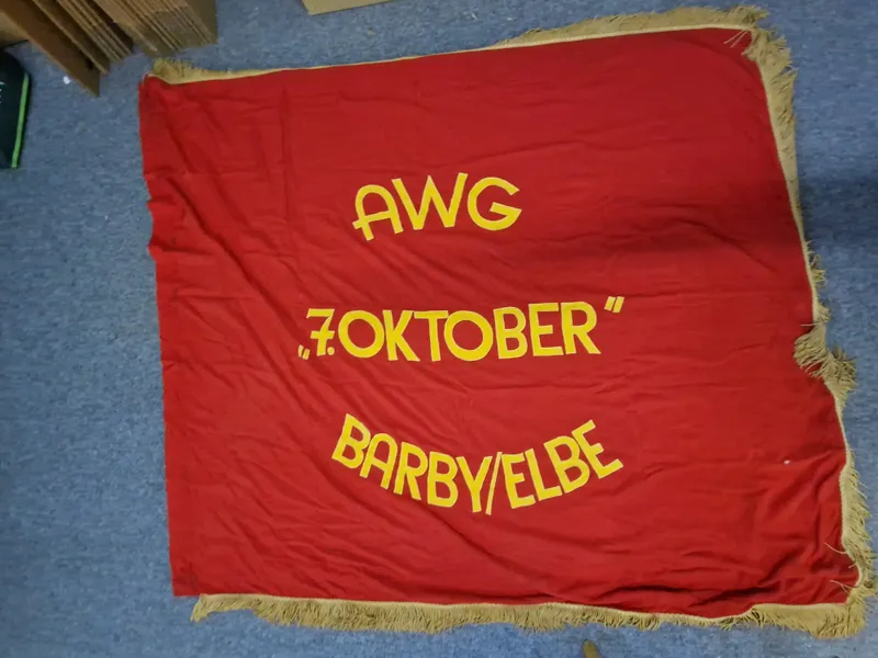 fane med teksten "AWG", "7. OKTOBER" og "BARBY/ELBE". 
AWG: står for "Arbeiterwohnungsbaugenossenschaft" (Arbejderboligbyggeriets Andelsforening), som var en type boligforening i DDR.